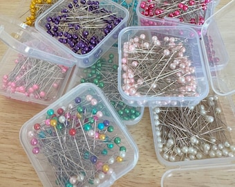 May include: A collection of clear plastic containers filled with sewing pins featuring round, pearl-like heads in various colors including pink, purple, yellow, green, white, and teal. The metal pins are arranged in multiple separate storage boxes.