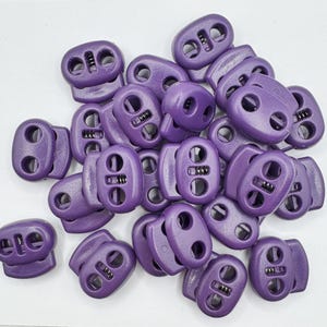 May include: A collection of purple cord locks, oval-shaped with two holes and a central mechanism for tightening cords. These plastic fasteners are designed for adjusting the fit of drawstrings or elastic cords. The image shows a pile of these small, functional components.