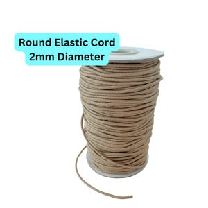 Może przedstawiać: Szpula beżowego, okrągłego sznurka elastycznego o średnicy 2 mm. Tekst "Round Elastic Cord 2mm Diameter" znajduje się w jasnoniebieskim prostokącie. Sznurek jest ciasno nawinięty na białą kartonową szpulę.