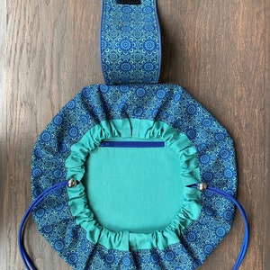 May include: A teal blue drawstring bag with a zippered pocket and a blue and white floral pattern. The bag has a blue strap with a velcro closure.