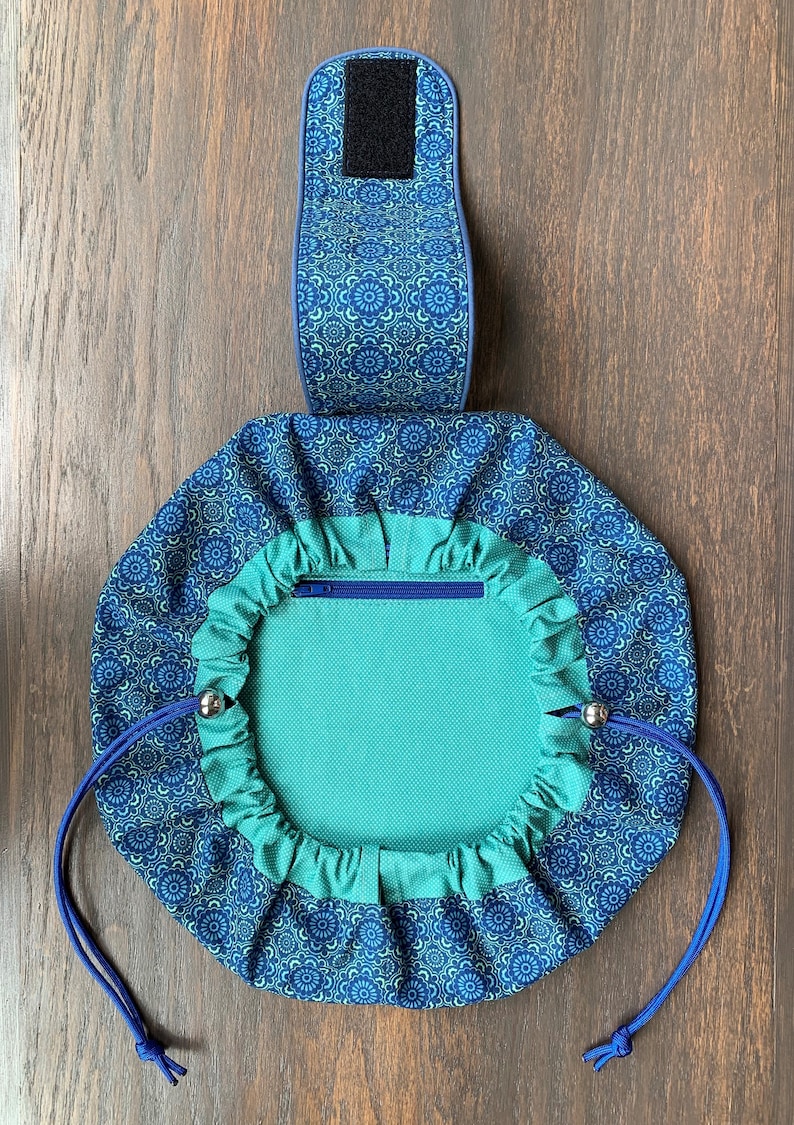 May include: A teal blue drawstring bag with a zippered pocket and a blue and white floral pattern. The bag has a blue strap with a velcro closure.
