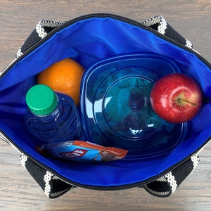 May include: A black and white striped lunch bag with a blue lining. The bag is open and contains a blue water bottle, an orange, a blue plastic container with a green salad, and a red apple.