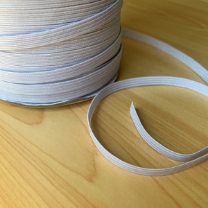 May include: A spool of white elastic ribbon, partially unrolled, sits on a light wood surface. The elastic is a pale gray color and appears to be tightly woven. The ribbon is likely used for sewing or crafting projects.