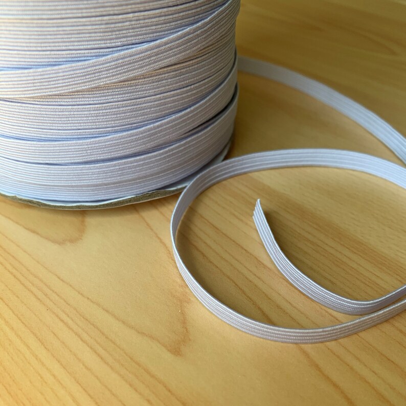 May include: A spool of white elastic ribbon, partially unrolled, sits on a light wood surface. The elastic is a pale gray color and appears to be tightly woven. The ribbon is likely used for sewing or crafting projects.