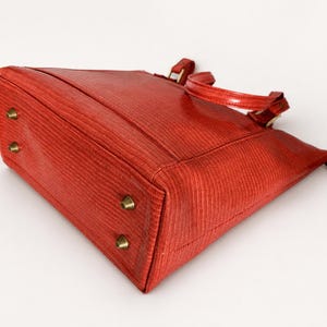 May include: A red handbag with a textured surface, featuring two top handles and four brass-toned metal feet on the bottom. The bag has a structured rectangular base and a zipper closure along the top edge, showcasing a consistent grain pattern.