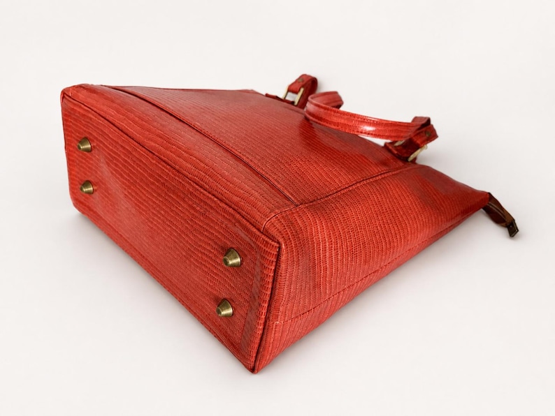May include: A red handbag with a textured surface, featuring two top handles and four brass-toned metal feet on the bottom. The bag has a structured rectangular base and a zipper closure along the top edge, showcasing a consistent grain pattern.