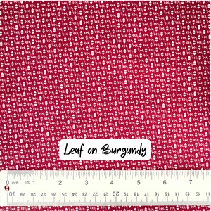 Op de afbeelding: Bordeaux stof met een herhalend wit bladerpatroon. Een liniaal met centimeter maten staat onderaan. De tekst "Leaf on Burgundy" is zichtbaar op de stof.