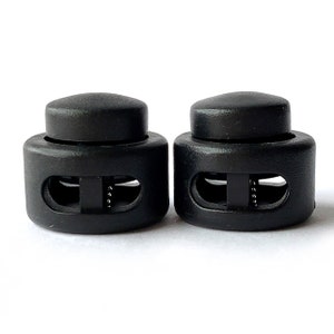 May include: Two black plastic cord locks with two holes each. The cord locks are designed to secure cords or straps.