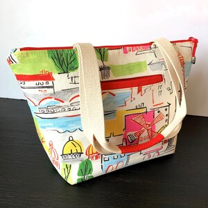 May include: A white canvas tote bag with red accents and a colorful city scene print. The bag has two cream-colored straps and a zipper closure.