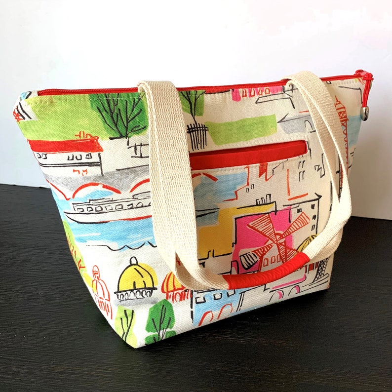 May include: A white canvas tote bag with red accents and a colorful city scene print. The bag has two cream-colored straps and a zipper closure.