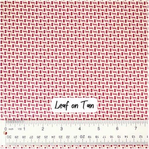 Op de afbeelding: Een stofstaal met een herhalend patroon van bordeauxrode bladachtige vormen op een tan achtergrond. De tekst "Leaf on Tan" staat op de stof. Een liniaal aan de onderkant toont de afmetingen in centimeters.