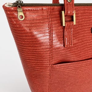 May include: A terracotta-colored tote bag with a textured, lizard-skin pattern. The bag features a top zipper closure with a brass-toned metal pull, matching brass-toned hardware at the strap attachments, and a visible exterior pocket.