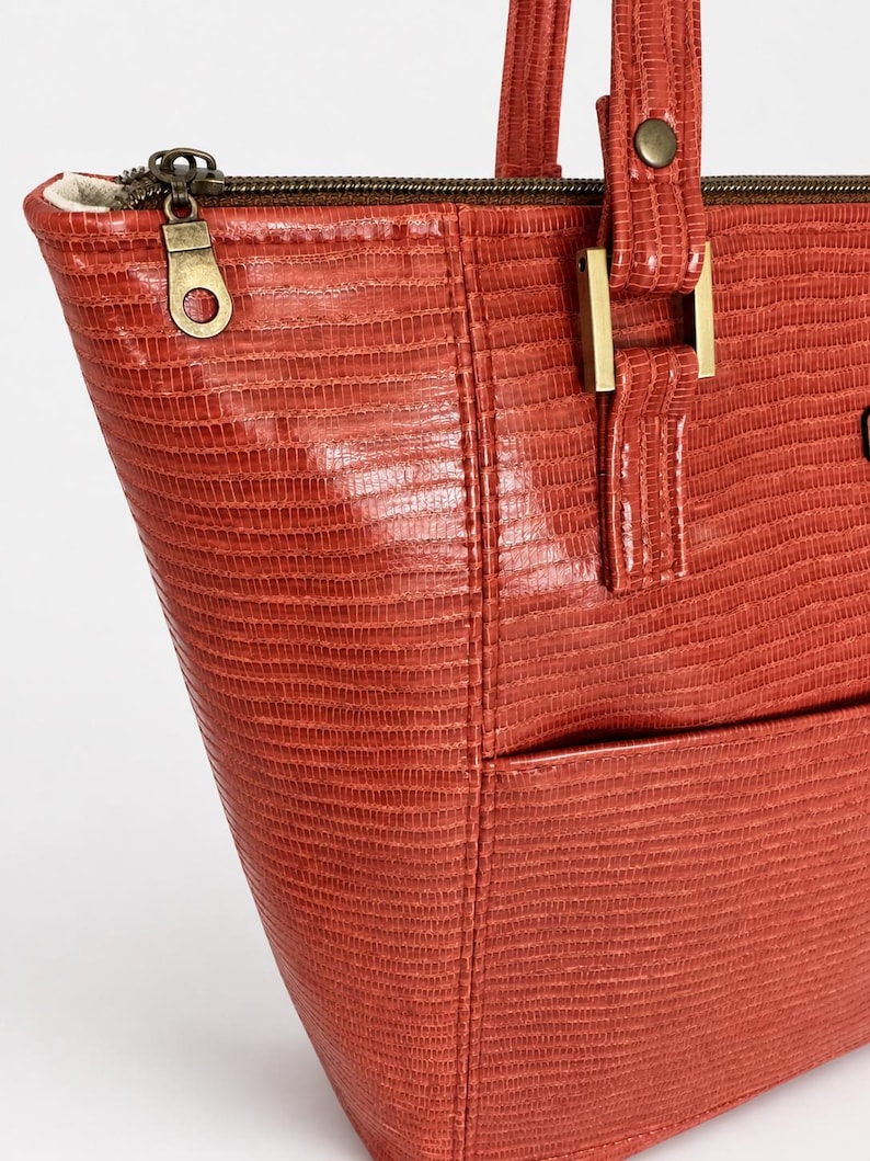 May include: A terracotta-colored tote bag with a textured, lizard-skin pattern. The bag features a top zipper closure with a brass-toned metal pull, matching brass-toned hardware at the strap attachments, and a visible exterior pocket.
