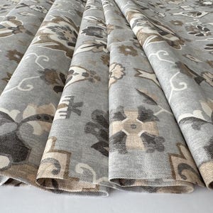 Op de afbeelding: Close-up van gevouwen stof met een bloemenpatroon. De stof is voornamelijk grijs met een herhalend ontwerp in tinten bruin, beige en wit. Het patroon bevat gestileerde bloemen en geometrische vormen.