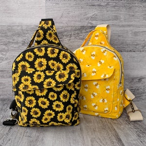Może przedstawiać: Dwa małe plecaki, jeden czarny z wzorem słonecznika, a drugi żółty z wzorem pszczoły.