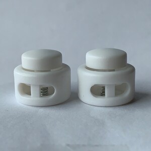 May include: Two white plastic cord locks with a spring mechanism. The cord locks have a round top and a rectangular base with two holes.
