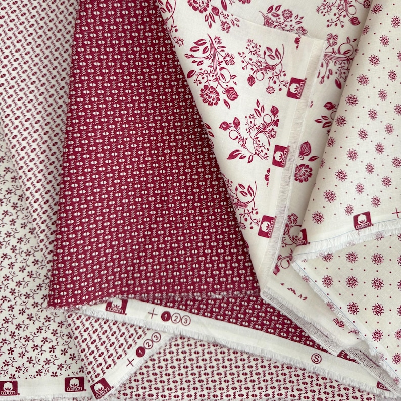 Op de afbeelding: Assortiment van katoenen stof met diverse bloemen- en geometrische patronen in bordeauxrood en wit. Ontwerpen omvatten kleine stippen, bloemtakjes en gestileerde bladvormen. De stof is gevouwen en gelaagd.