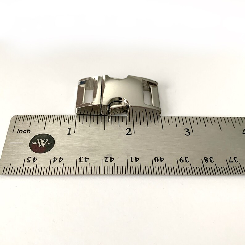 May include: Silver metal slider buckle measuring 1 inch in length. The buckle is shown next to a ruler for size reference.