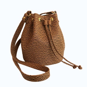 Può includere: Una borsa a secchiello in pelle marrone con chiusura a coulisse e una tracolla lunga. La borsa ha una trama testurizzata, simile a quella del cuoio di struzzo.