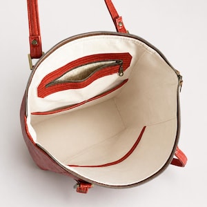 May include: A red tote bag with a textured exterior and two shoulder straps. The interior is lined with off-white fabric and features a zippered pocket with a metallic gold accent, along with an open slip pocket on the opposite side.