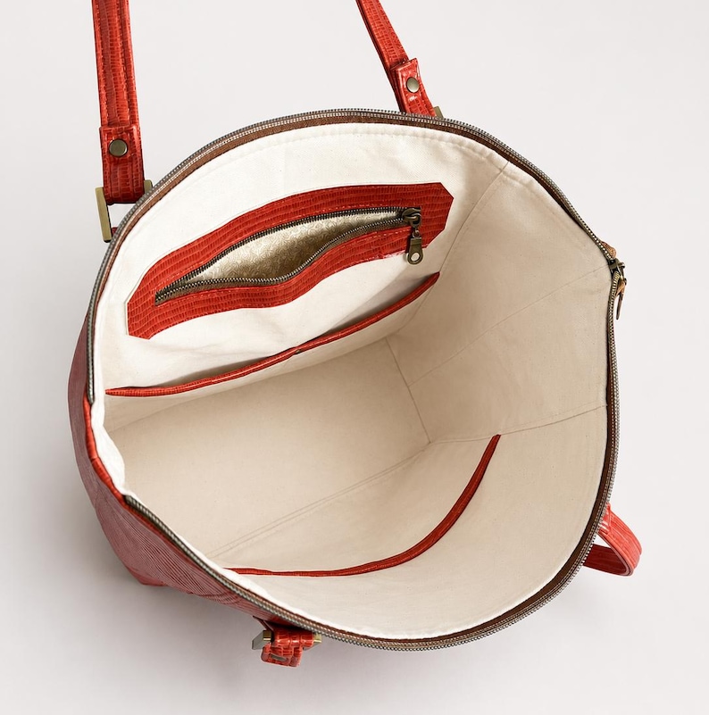 May include: A red tote bag with a textured exterior and two shoulder straps. The interior is lined with off-white fabric and features a zippered pocket with a metallic gold accent, along with an open slip pocket on the opposite side.