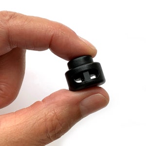 May include: A black plastic toggle with a small hole in the center. The toggle is designed to be used for adjusting the length of cords or straps.