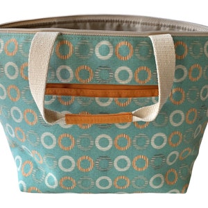 May include: A teal canvas tote bag with cream-colored handles and a zipper closure. The bag has a repeating pattern of orange and white circles on a teal background.