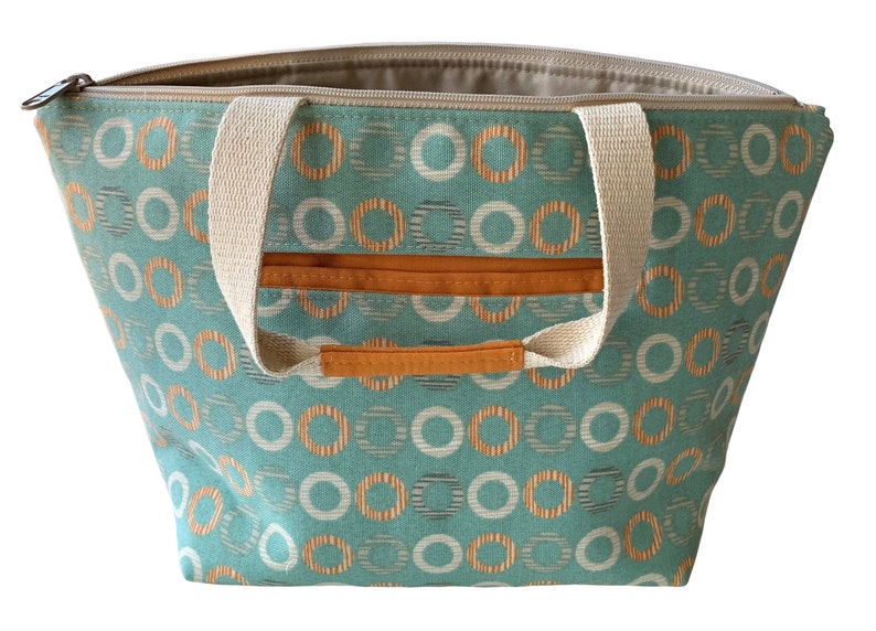 May include: A teal canvas tote bag with cream-colored handles and a zipper closure. The bag has a repeating pattern of orange and white circles on a teal background.