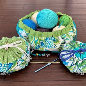May include: Three green and blue floral drawstring bags, labeled "Large", "Medium", and "Small". The large bag is filled with colorful yarn balls. Two crochet hooks are on the table.