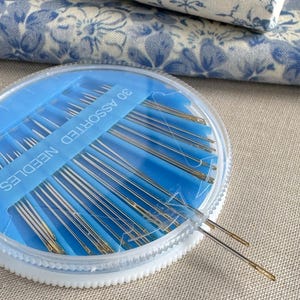 Puede incluir: Un recipiente de plástico azul que contiene 30 agujas surtidas. Las agujas son plateadas con ojales dorados. El recipiente es redondo con una tapa transparente y un borde blanco estriado. Tela floral azul en el fondo.