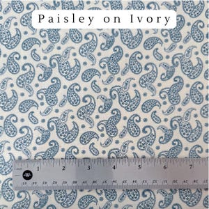May include: Ivory fabric with a blue paisley pattern. The fabric has a repeating design of teardrop-shaped paisley motifs. The text "Paisley on Ivory" is at the top. A ruler is at the bottom, showing inches.