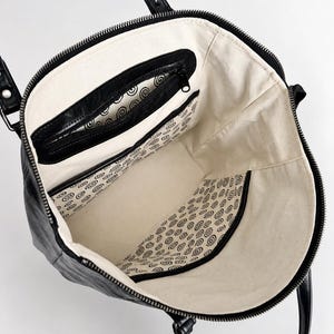 May include: A black tote bag with a top zipper closure and two shoulder straps. The interior is lined with off-white fabric featuring a repeating black spiral pattern. It includes a black zippered pocket and an open slip pocket inside.