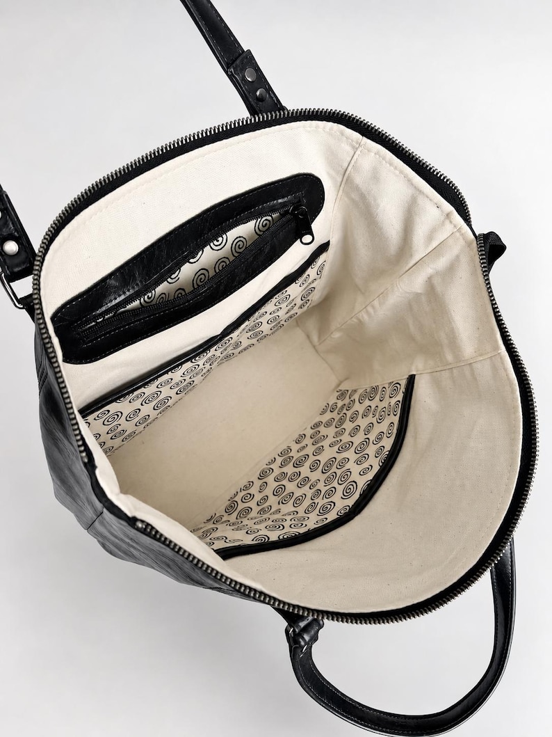 May include: A black tote bag with a top zipper closure and two shoulder straps. The interior is lined with off-white fabric featuring a repeating black spiral pattern. It includes a black zippered pocket and an open slip pocket inside.