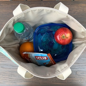 May include: A blue and white patterned tote bag with a zipper closure and two handles. The bag is open and contains a clear water bottle, a blue plastic container with a green salad, a red apple, an orange, and a brown and orange Cliff Bar wrapper.