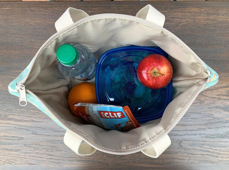 May include: A blue and white patterned tote bag with a zipper closure and two handles. The bag is open and contains a clear water bottle, a blue plastic container with a green salad, a red apple, an orange, and a brown and orange Cliff Bar wrapper.