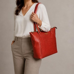 May include: A red tote bag with a textured, horizontal-lined surface pattern. The bag features two long shoulder straps attached with rectangular hardware, a top zipper closure, and a small rectangular brand label on the front exterior panel.