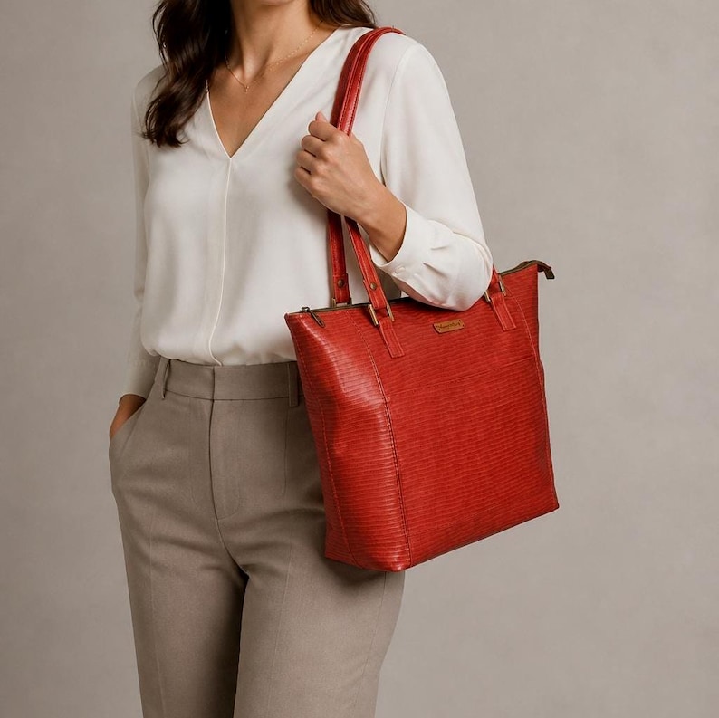 May include: A red tote bag with a textured, horizontal-lined surface pattern. The bag features two long shoulder straps attached with rectangular hardware, a top zipper closure, and a small rectangular brand label on the front exterior panel.