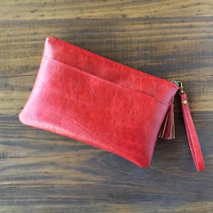 May include: A bright red rectangular clutch bag with a textured surface, featuring a horizontal front pocket, a matching red wrist strap, and a decorative tassel attached to the metal zip hardware on the side of the accessory.
