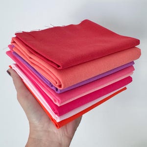以下が含まれることがあります： 深紅、コーラル、紫、ライトピンク、マゼンタ、鮮やかなオレンジといった暖色系のグラデーションで折り畳まれた布の正方形の束。テキスタイルには目に見える織り目があり、上部の折り目には切りっぱなしの端とほつれた糸が見られます。