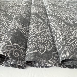 Puede incluir: Primer plano de tela gris doblada con un estampado floral y paisley blanco. La tela tiene un aspecto texturizado y probablemente se utiliza para la decoración del hogar o textiles. El diseño presenta elementos botánicos detallados.