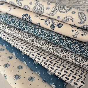 May include: A stack of folded fabric with various blue and white floral and paisley patterns. The fabrics have different textures and shades of blue, creating a visually appealing display of textiles. The patterns include small flowers, vines, and classic paisley designs.