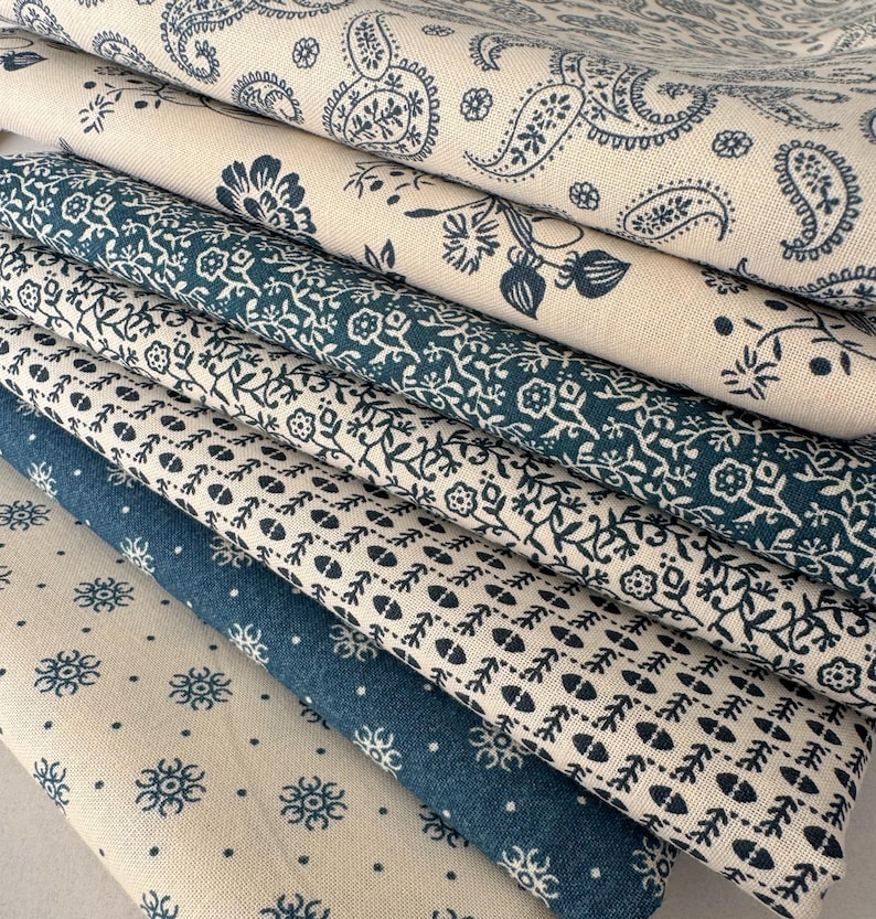 May include: A stack of folded fabric with various blue and white floral and paisley patterns. The fabrics have different textures and shades of blue, creating a visually appealing display of textiles. The patterns include small flowers, vines, and classic paisley designs.