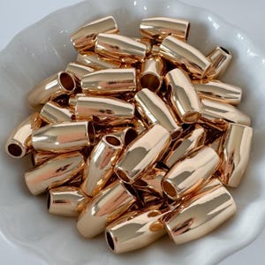 May include: A collection of shiny, gold-colored barrel-shaped beads. Each bead has a smooth, polished surface and a cylindrical hole at each end. The beads are arranged in a white, shallow bowl.