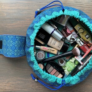 May include: A teal and blue round makeup bag with a drawstring closure. The bag is open and filled with various makeup products, including foundation, concealer, eyeshadow palettes, mascara, eyeliner, and brushes.
