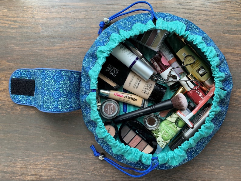 May include: A teal and blue round makeup bag with a drawstring closure. The bag is open and filled with various makeup products, including foundation, concealer, eyeshadow palettes, mascara, eyeliner, and brushes.