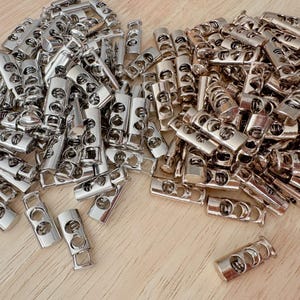 May include: A collection of shiny, silver metal cord locks. These cylindrical cord locks have two holes and a rectangular base. The cord locks are scattered on a light brown wooden surface.