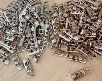 Op de afbeelding: Een verzameling glanzende, zilveren metalen koordstoppers. Deze cilindrische koordstoppers hebben twee gaten en een rechthoekige basis. De koordstoppers liggen verspreid op een lichtbruin houten oppervlak.