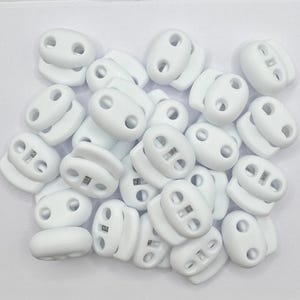 May include: A collection of white cord locks, oval-shaped with two holes and a spring mechanism. These plastic cord locks are designed to secure and adjust cords, commonly used in apparel and accessories.