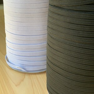 May include: Two spools of elastic ribbon. One spool is white, and the other is dark brown. The elastic is tightly wound on cardboard spools. The elastic is likely used for sewing or crafting projects.