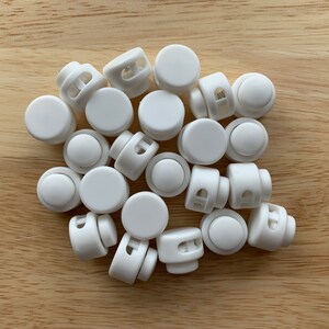 May include: A pile of white plastic toggle clasps, also known as slider buckles, for use in sewing and crafting projects.